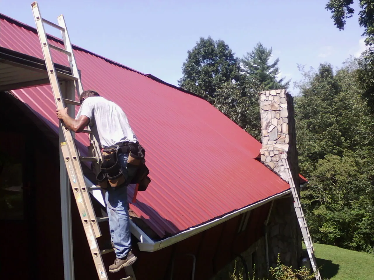 Roofer on a ladder beside a red metal roof and stone chimney — Soffit & Fascia Repair by Roofing Brothers in Sherrodsville
